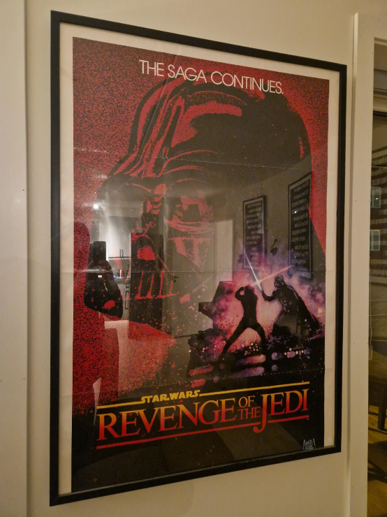 Framed "Revenge of the Jedi" movie poster featuring Darth Vader's helmet and two figures with lightsabers beneath the text "The Saga Continues"