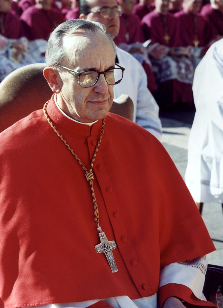 Cardinal Bergoglio in formal attire with glasses and a cross necklace sits during a ceremony, surrounded by similarly dressed clergy members