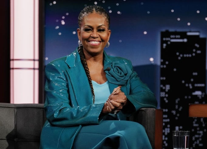 Person seated on a talk show set, wearing a stylish suit with metallic shoes and braided hairstyle, smiling warmly