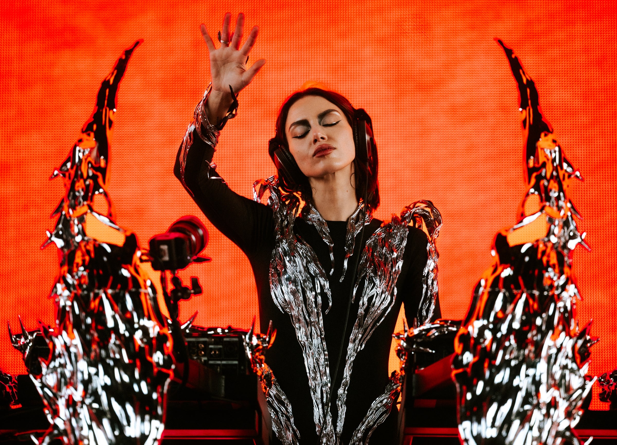 Musician performing energetically on stage in futuristic outfit with dramatic metallic accents, hand raised, vivid background contrasts performance