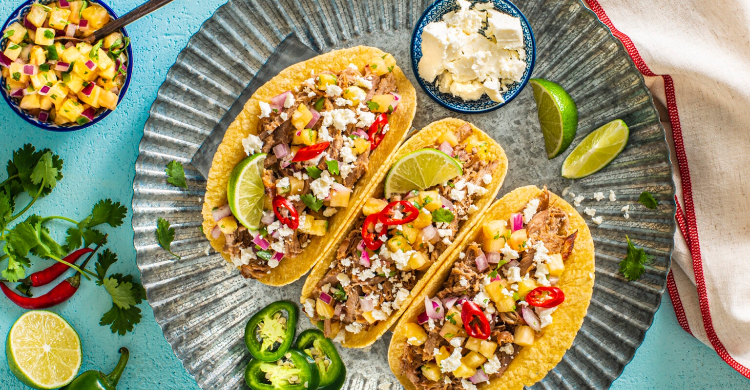 Three tacos on a metal plate, filled with meat, diced pineapple, onions, cotija cheese, and red chili slices, garnished with lime and cilantro