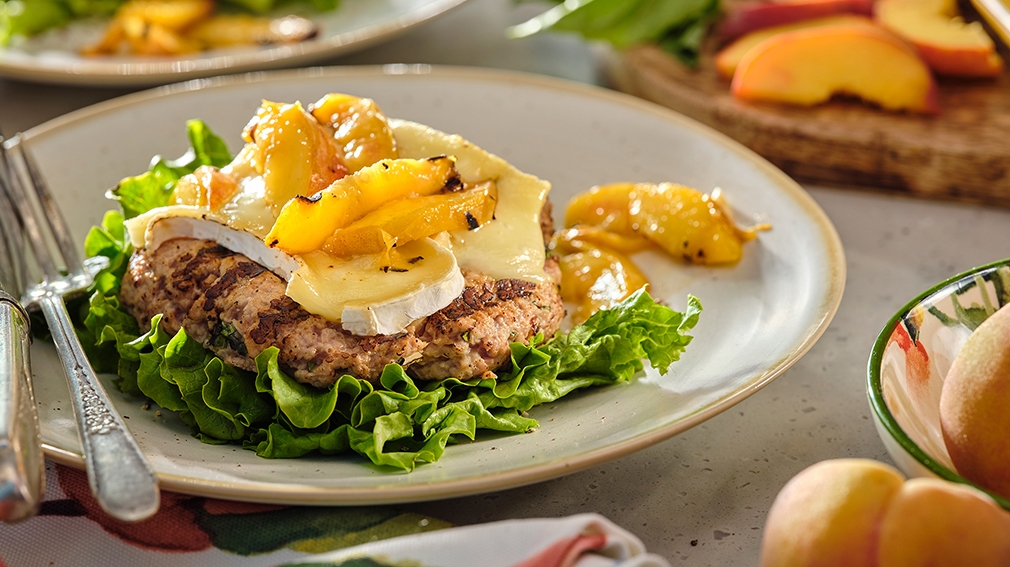 A gourmet burger patty on lettuce, topped with brie and grilled peaches on a plate, surrounded by fresh peach slices