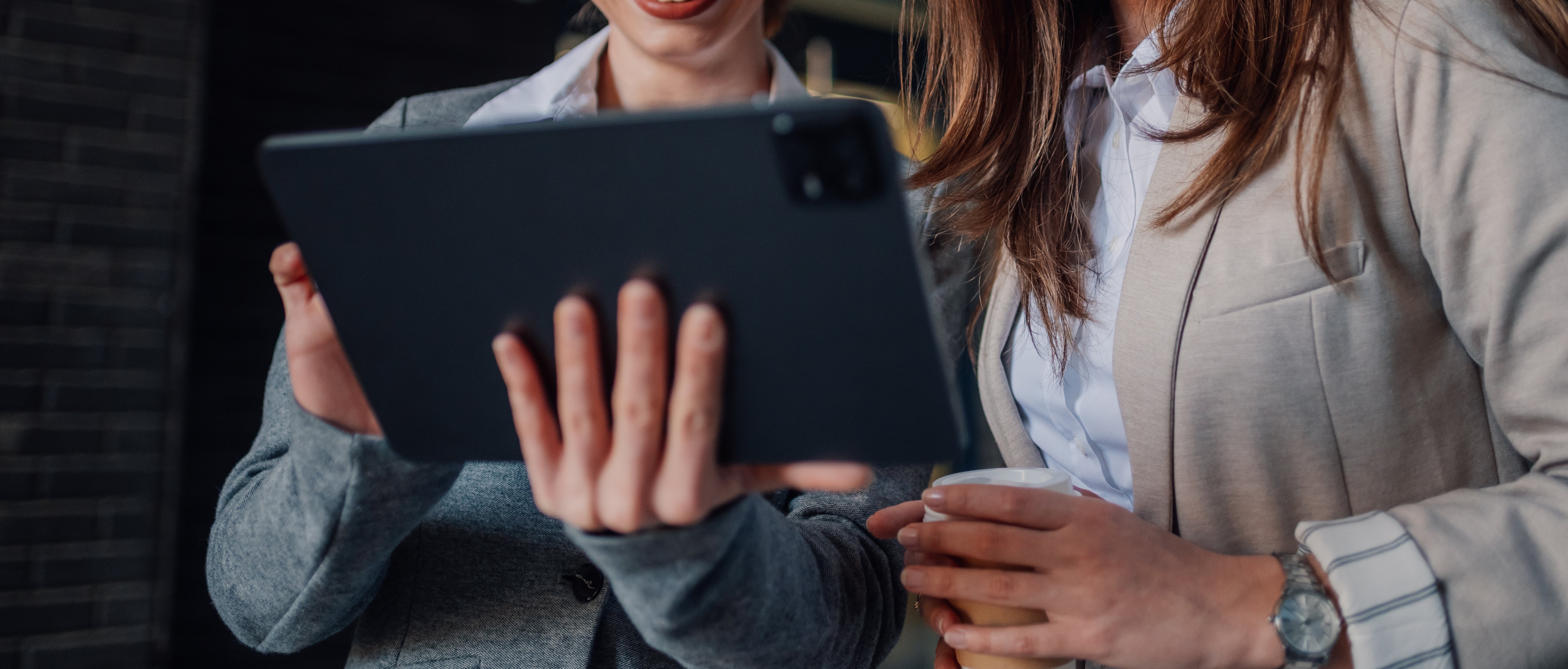 Two women happily look at a tablet together, one holding a coffee cup, both wearing business attire