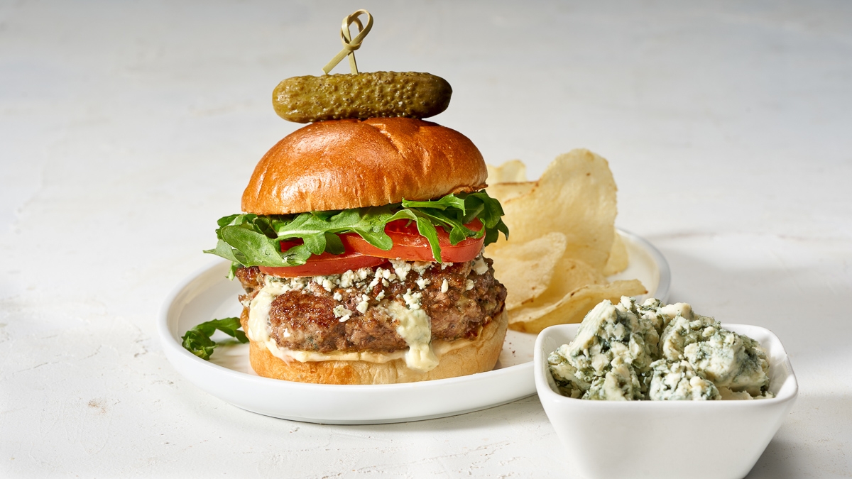 Gourmet burger with lettuce, tomato, and cheese, topped with a pickle on a skewer, served with chips and a side of crumbled blue cheese