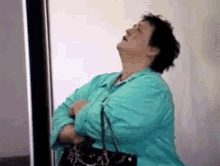 Person in a teal shirt sighs and looks upward while waiting by an elevator, holding a bag