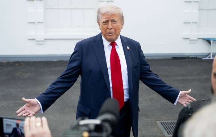 Person standing outdoors in a suit and red tie, gesturing with arms open, speaking to reporters