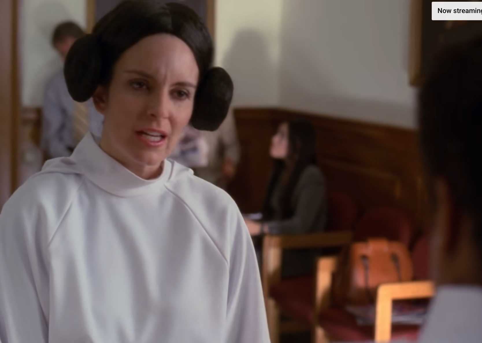 The image shows a person in a courtroom setting wearing a white outfit with hair styled in two buns. Another person is in the foreground