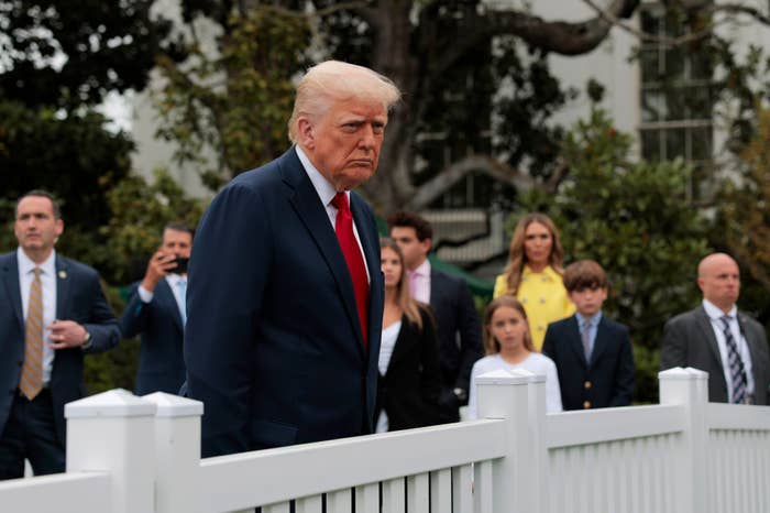A group of people, including a man in a suit, are gathered outside near a white fence, with trees in the background