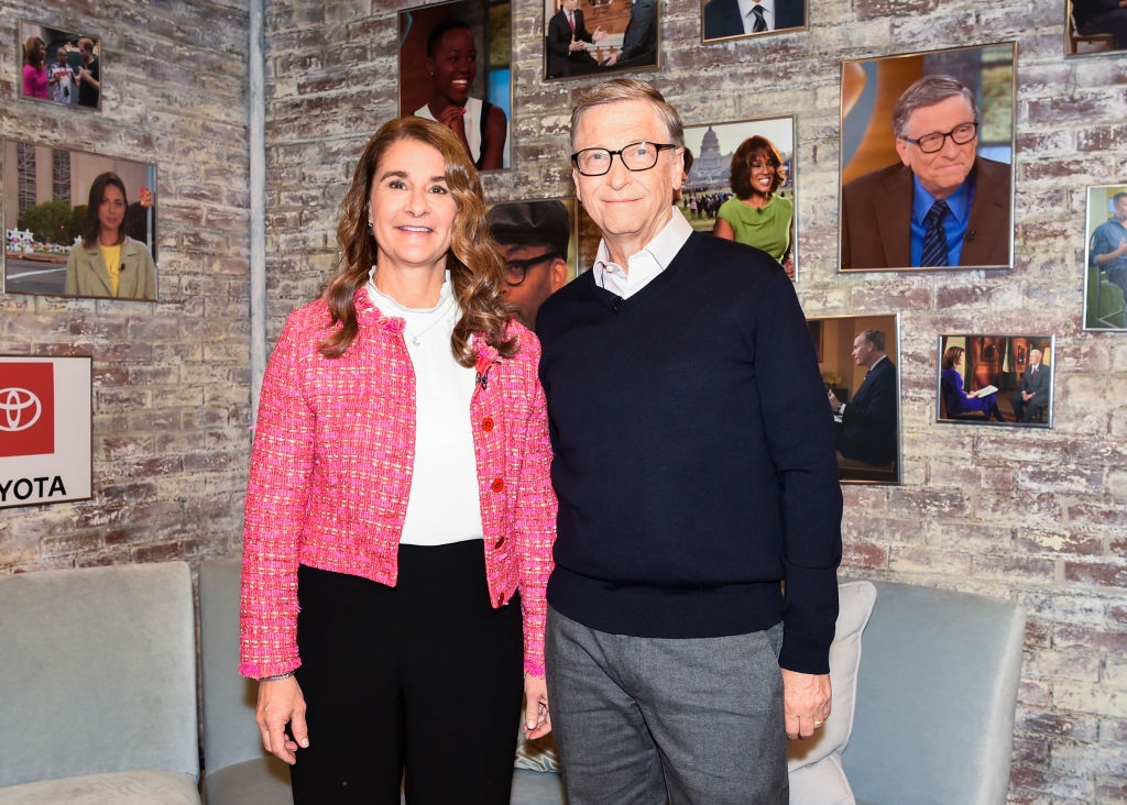 Two individuals posing in a living room set surrounded by framed photos. One wears a tweed jacket, and the other wears a sweater over a collared shirt