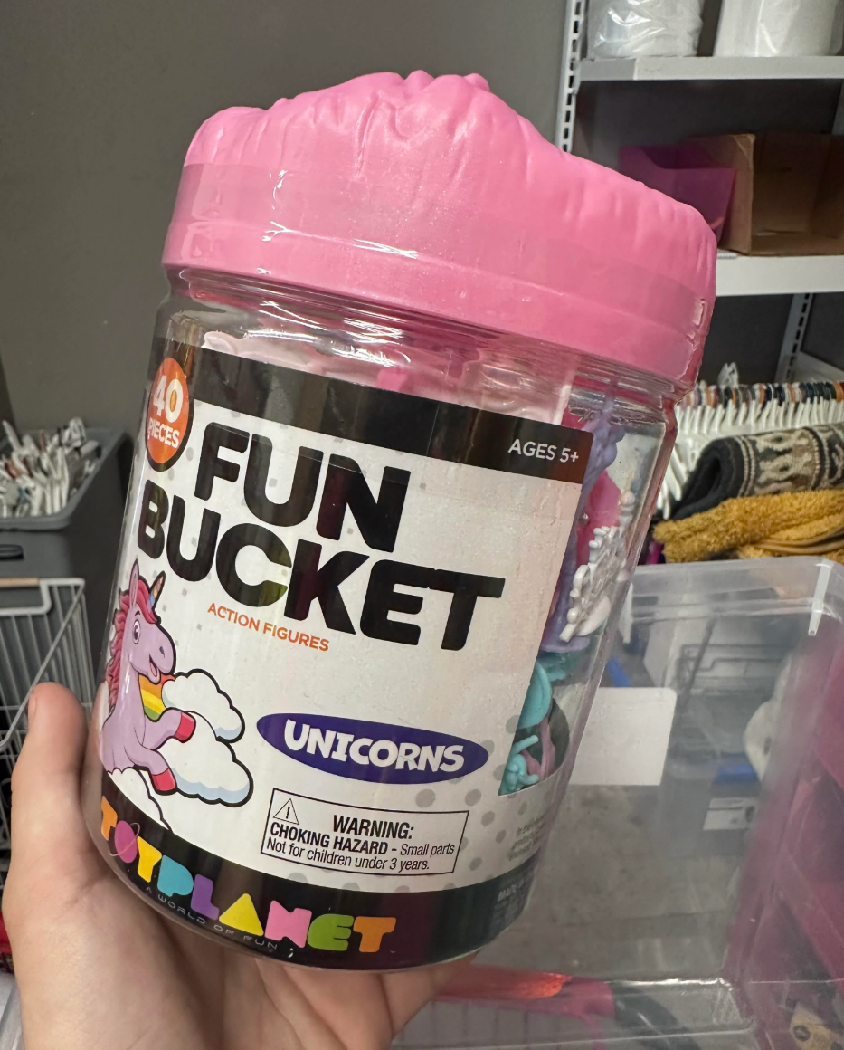 A large jar labeled "40 Fun Bucket Action Figures: Unicorns" is held in a store. Warning: Choking hazard for small parts, not for children under 3