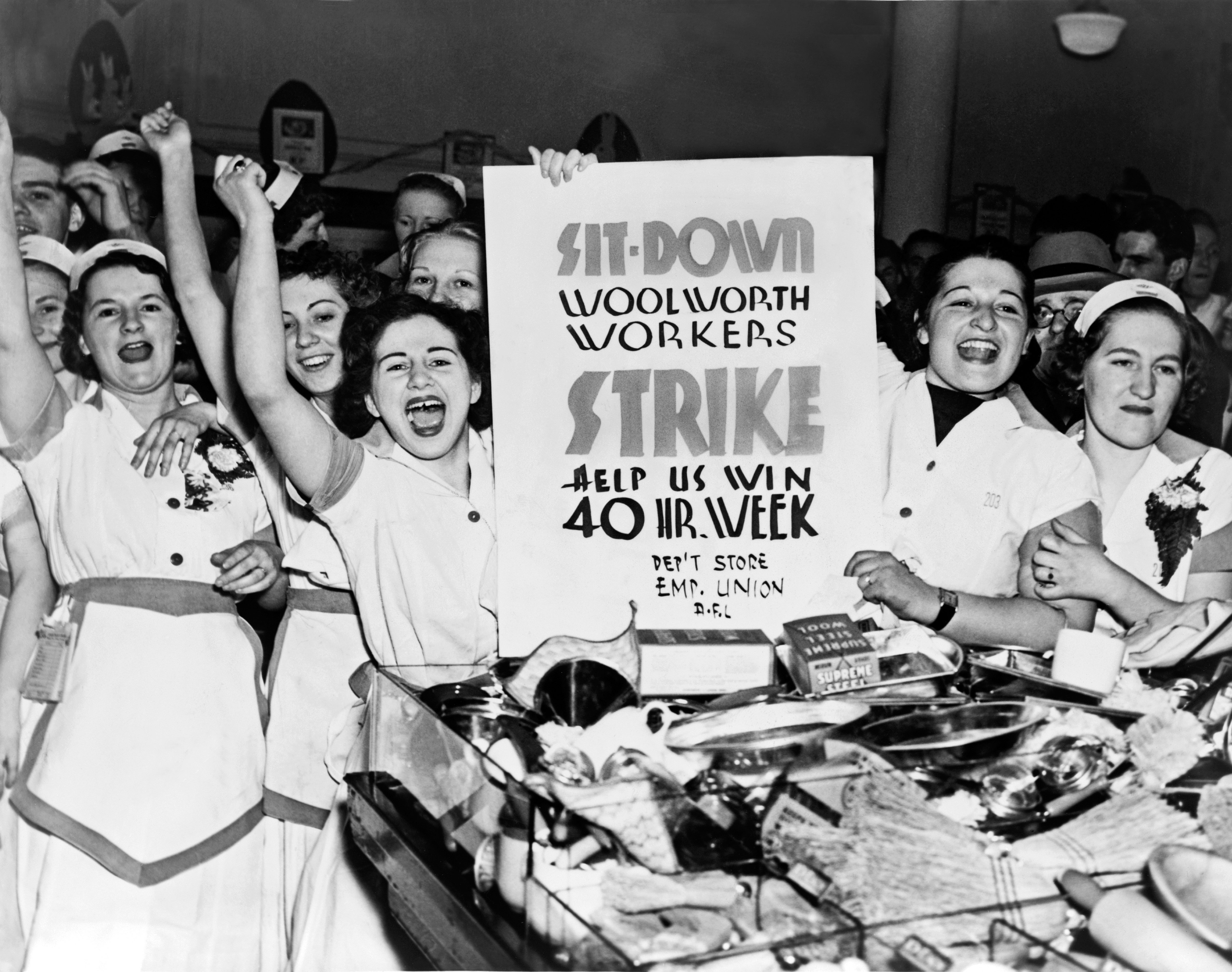Group of women hold a "Sit-Down Woolworth Workers Strike" sign, celebrating in a crowded store during a labor protest for a 40-hour workweek