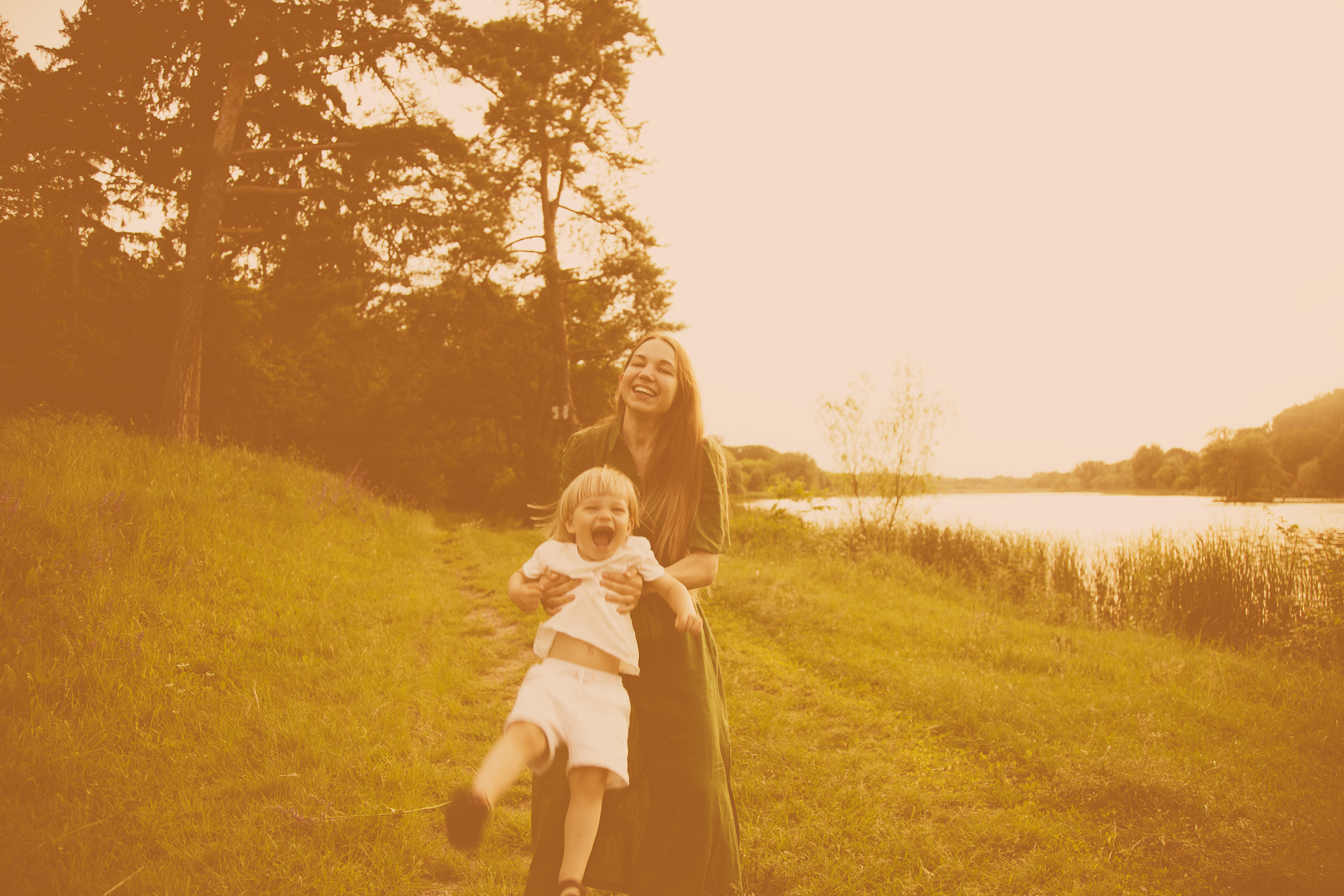 Woman joyfully spins child in park near water, both smiling broadly