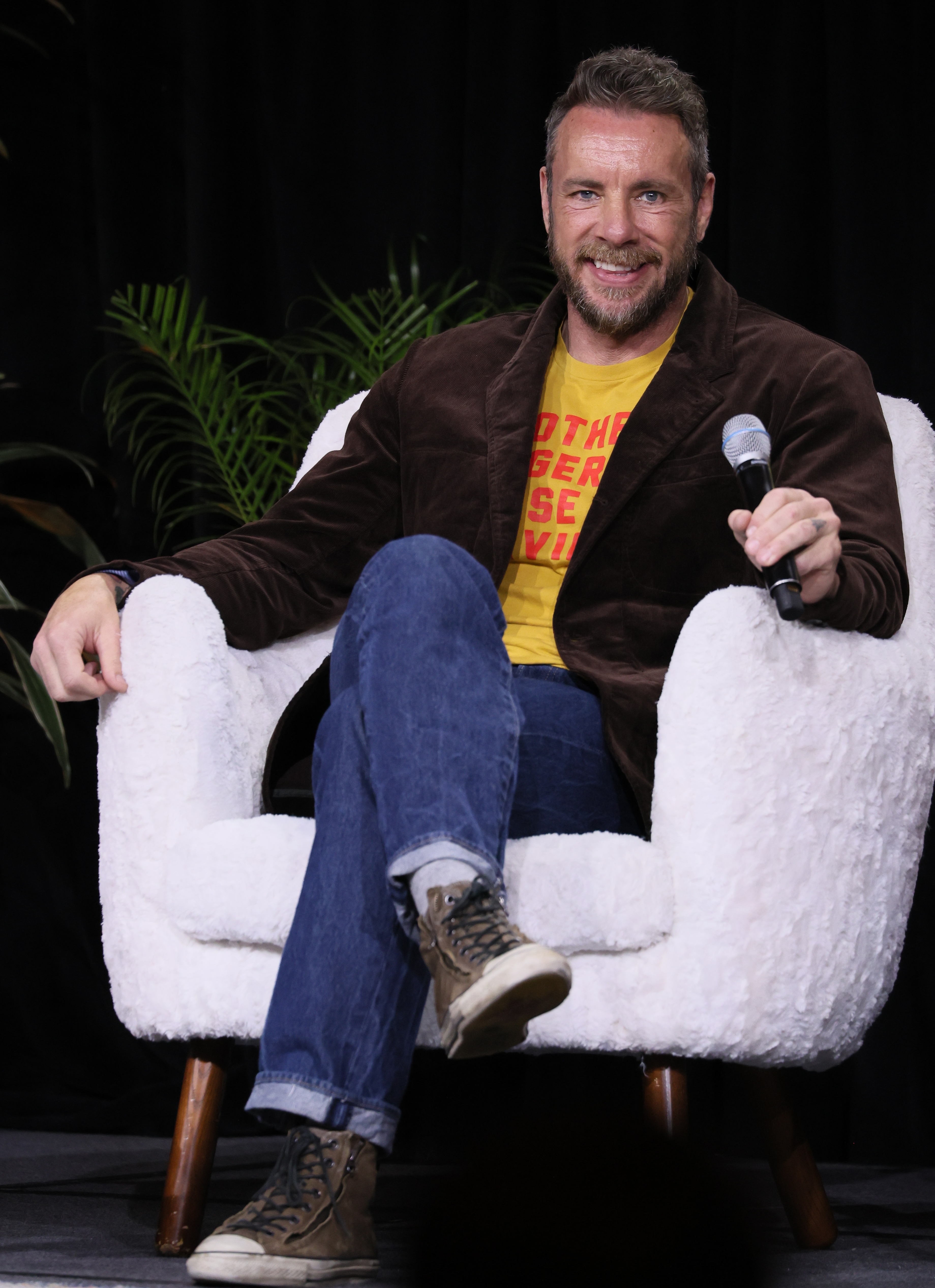 Person seated in a fluffy chair, wearing a brown jacket, yellow graphic tee, jeans, and sneakers, holding a microphone and smiling