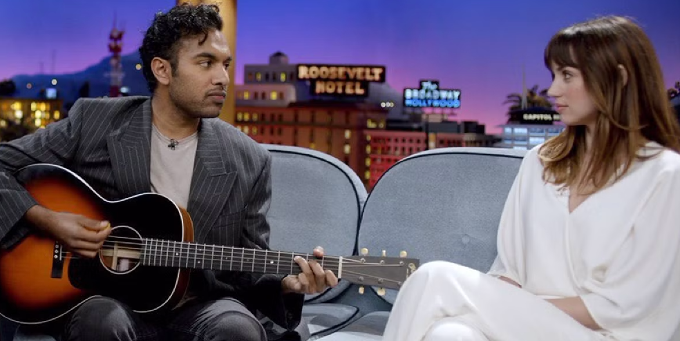 Man in pinstripe suit playing guitar, sitting next to woman in white outfit on a talk show set with neon signs in the background