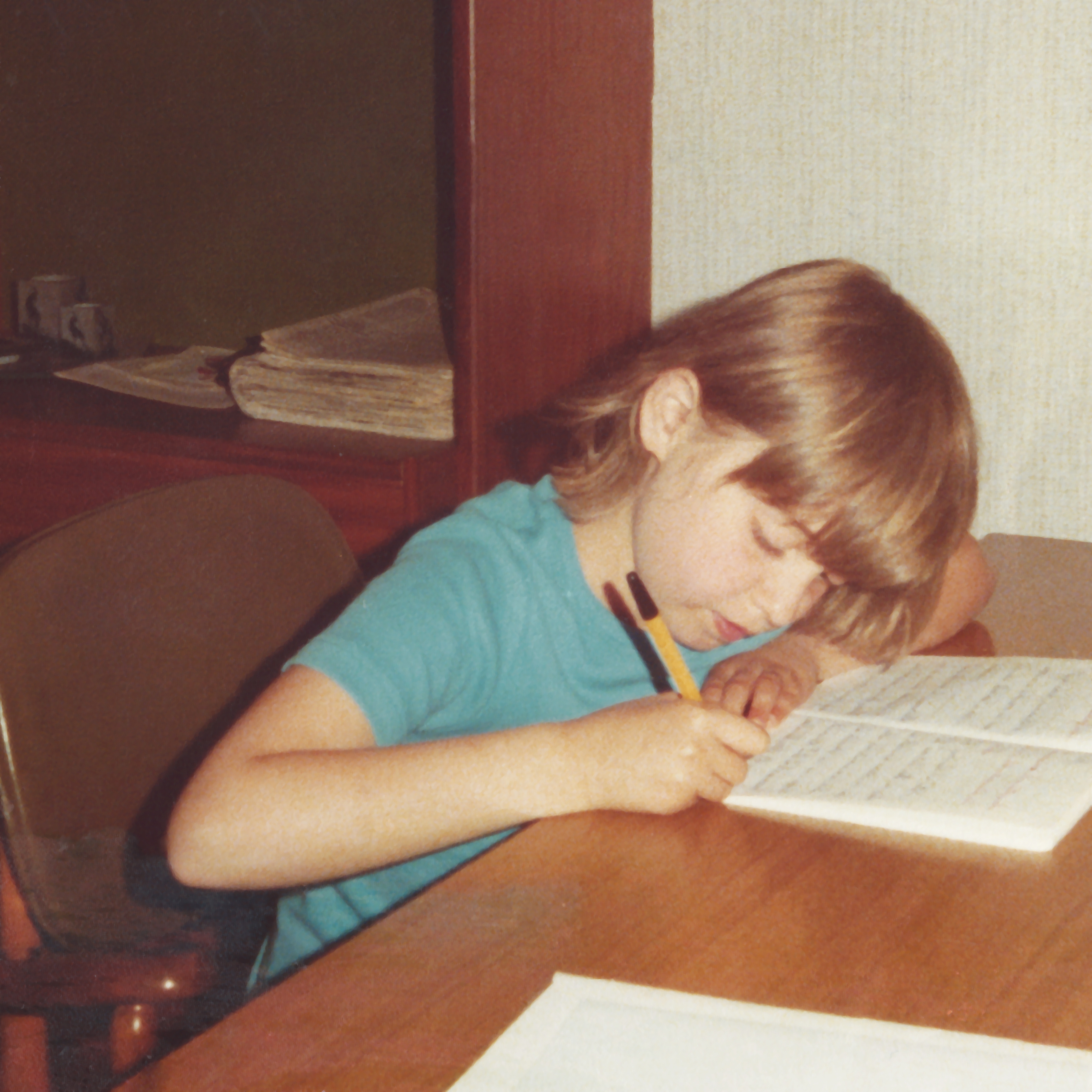 Child writing in a notebook at a wooden table, focused and seated on a chair. The room has books and papers on a shelf in the background