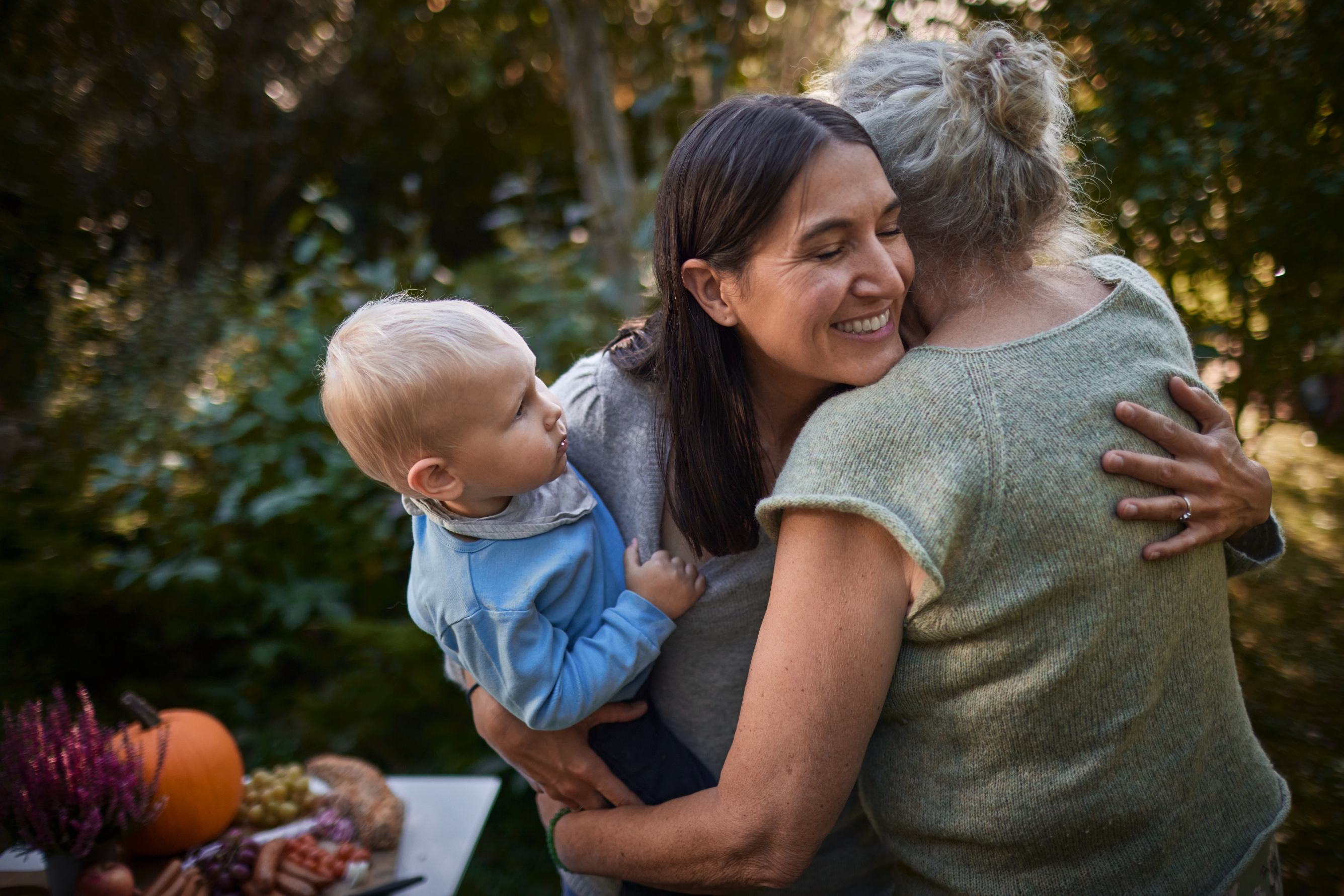 Woman holding a toddler hugs an older woman in a garden setting, with a table of autumnal decorations nearby