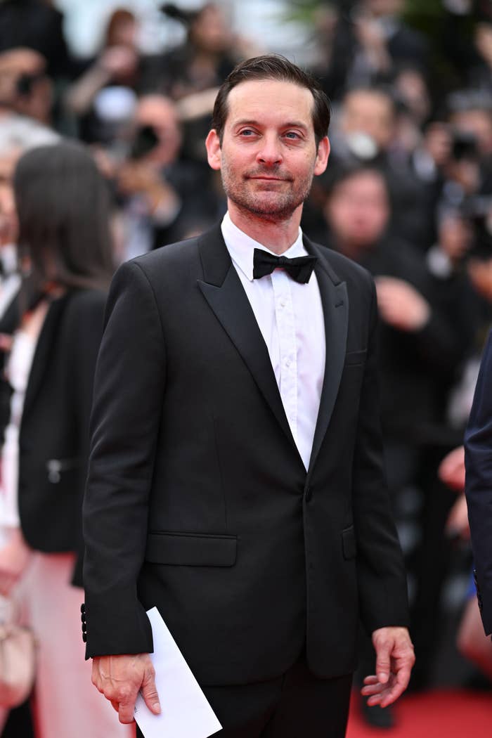 A person in a classic black tuxedo with a bow tie stands on a red carpet, holding a paper, amidst a crowd of people