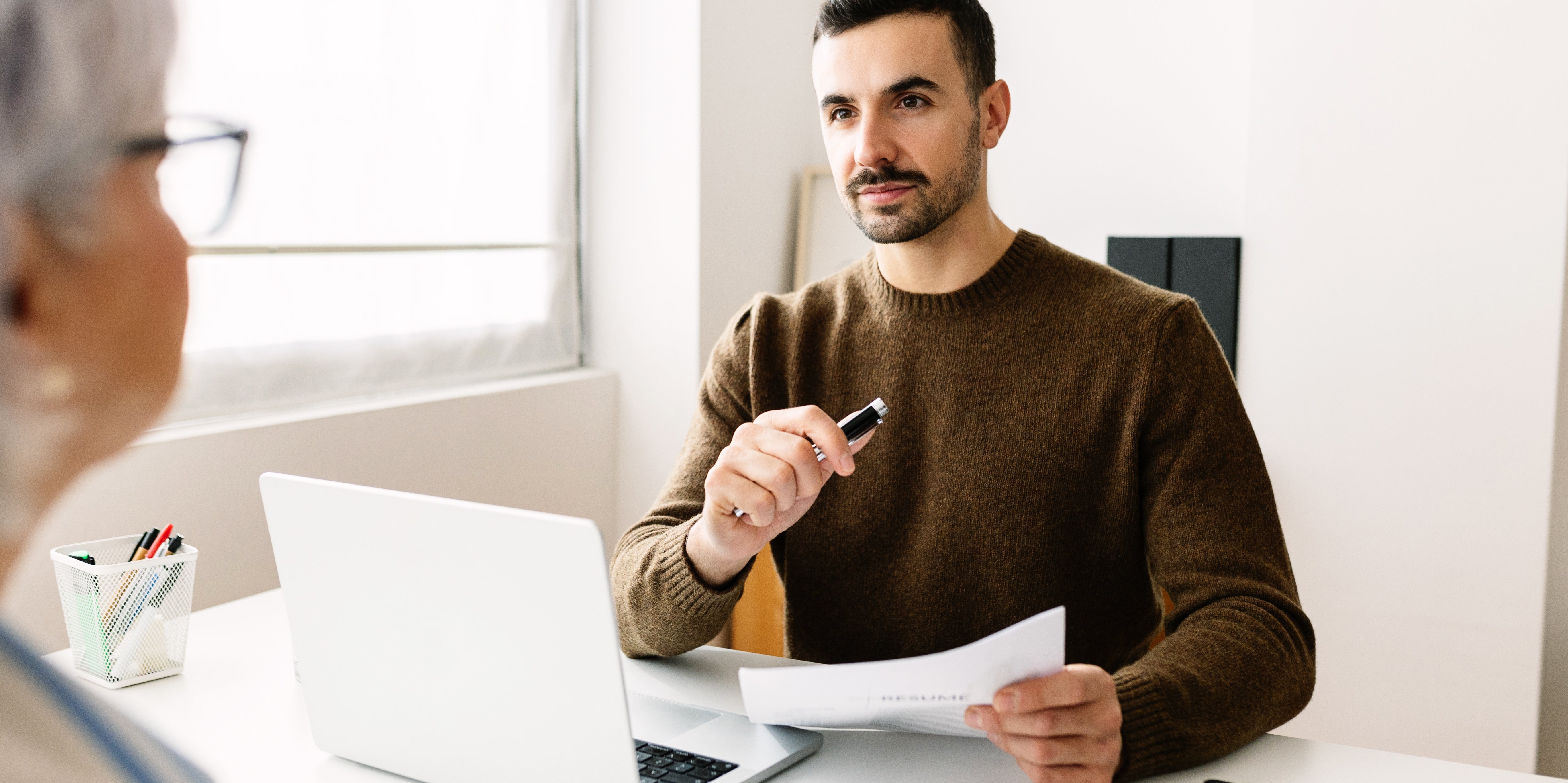 Person holding a pen, sitting at a desk with a laptop, paper, and phone, talking to someone out of view. Office setting
