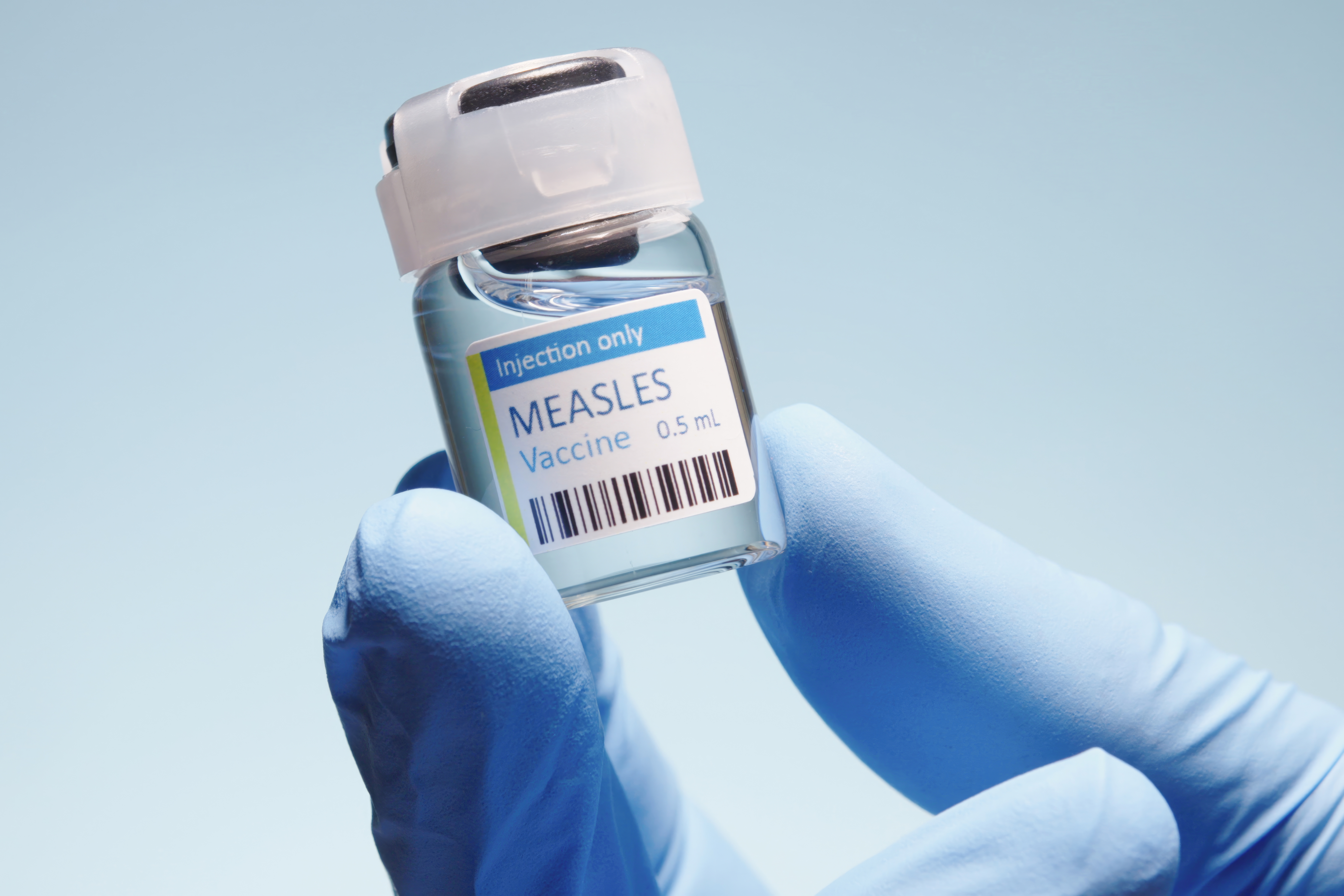 Hand holding a small vial labeled "Measles Vaccine 0.5 mL," emphasizing immunization. Close-up with a gloved hand, symbolizing medical safety