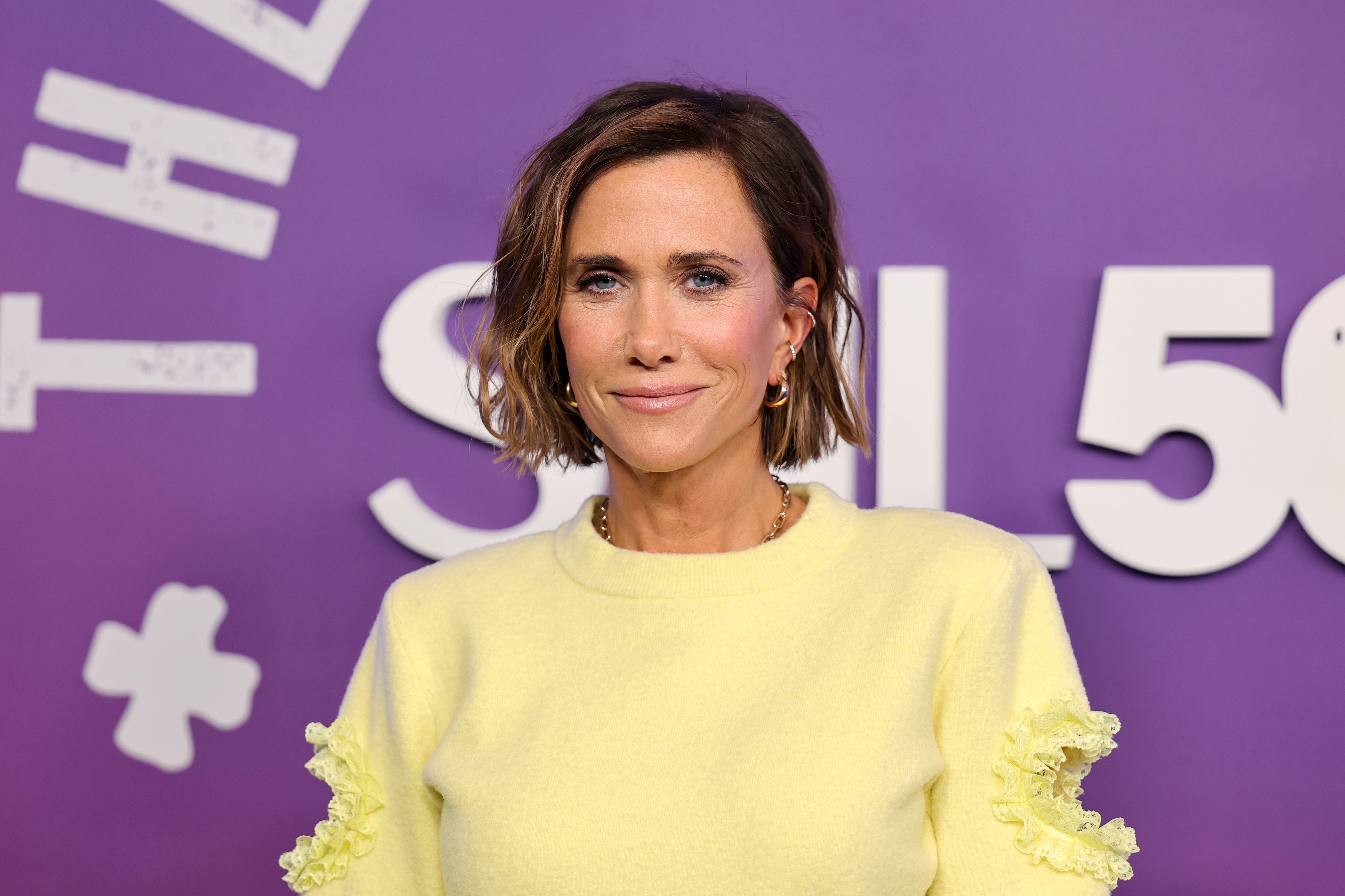 Person in a stylish outfit with lace detailing, standing against a backdrop featuring "SNL" text, attending a TV and movies-related event