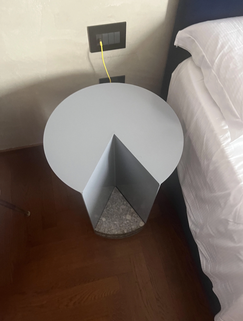 Uniquely designed bedside table with a missing pie-shaped section, placed next to a bed with white bedding