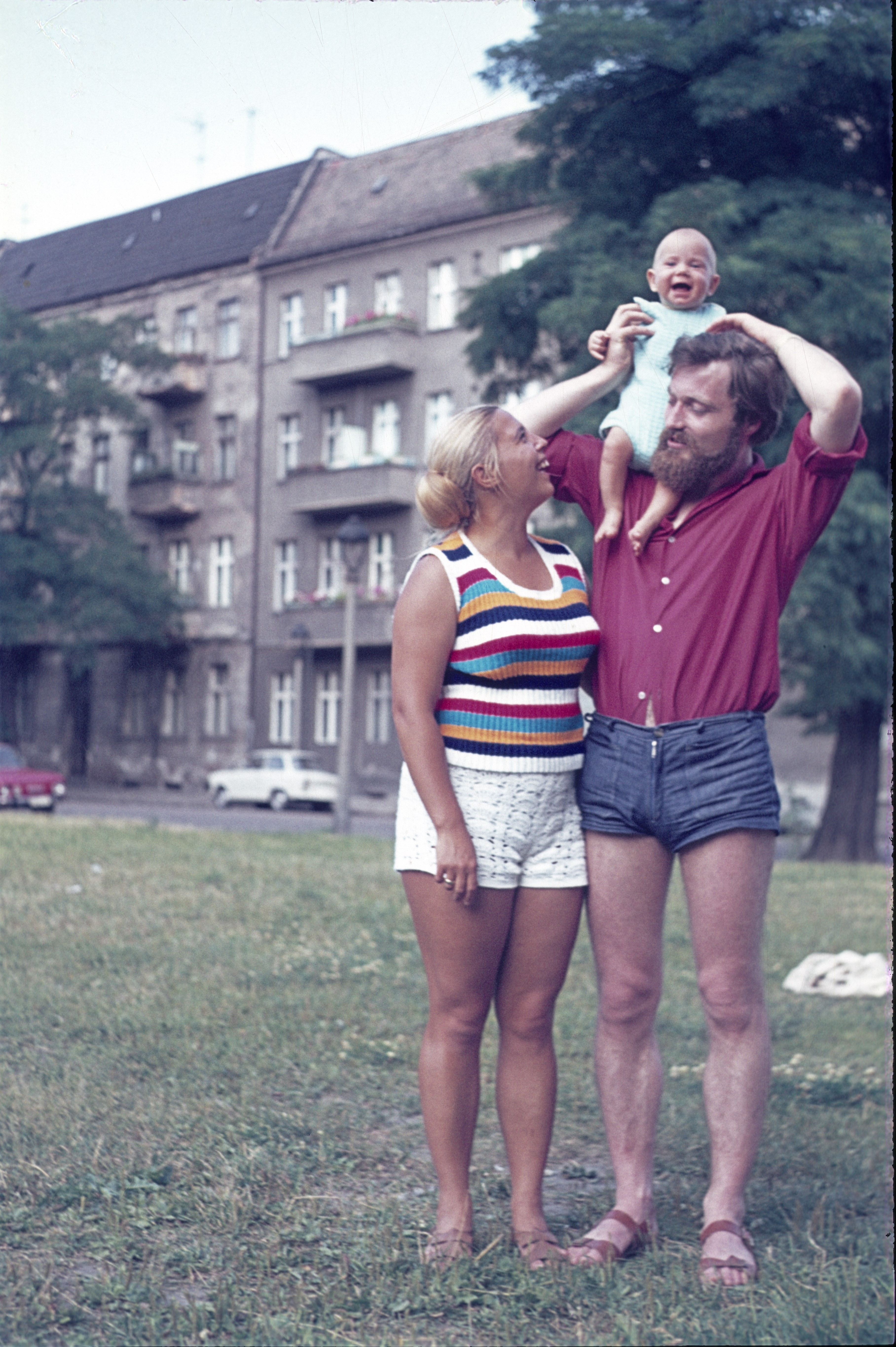 A family stands outdoors; a man in shorts holds a laughing baby on his shoulders, while a woman in a sleeveless top and shorts looks on smiling