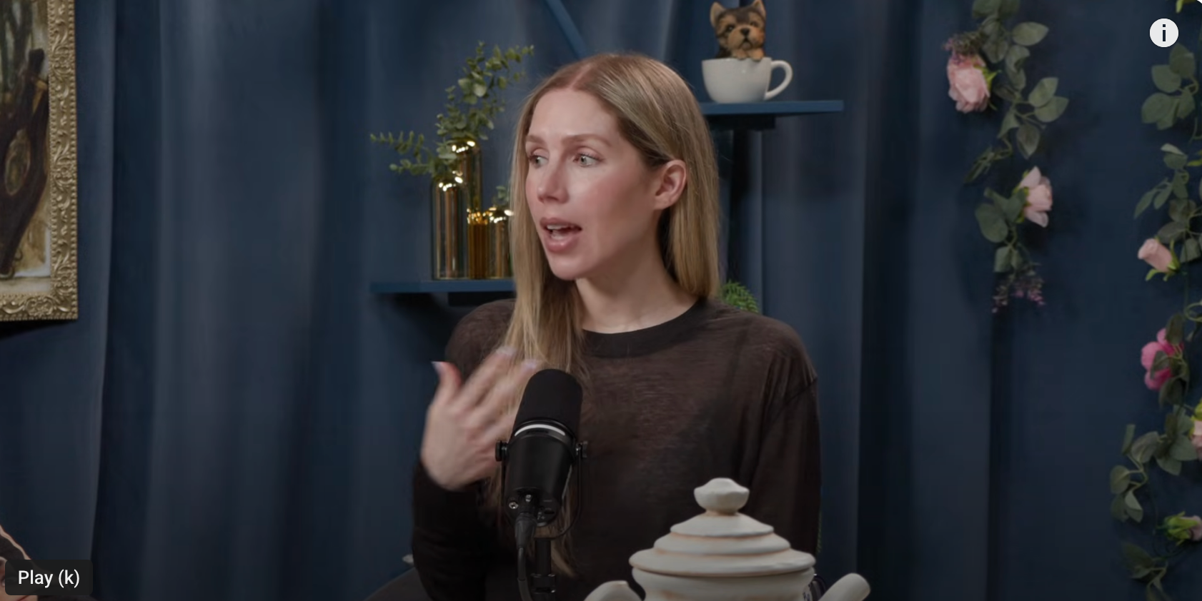 Person with long hair speaking into a microphone in a studio decorated with plants and teapots