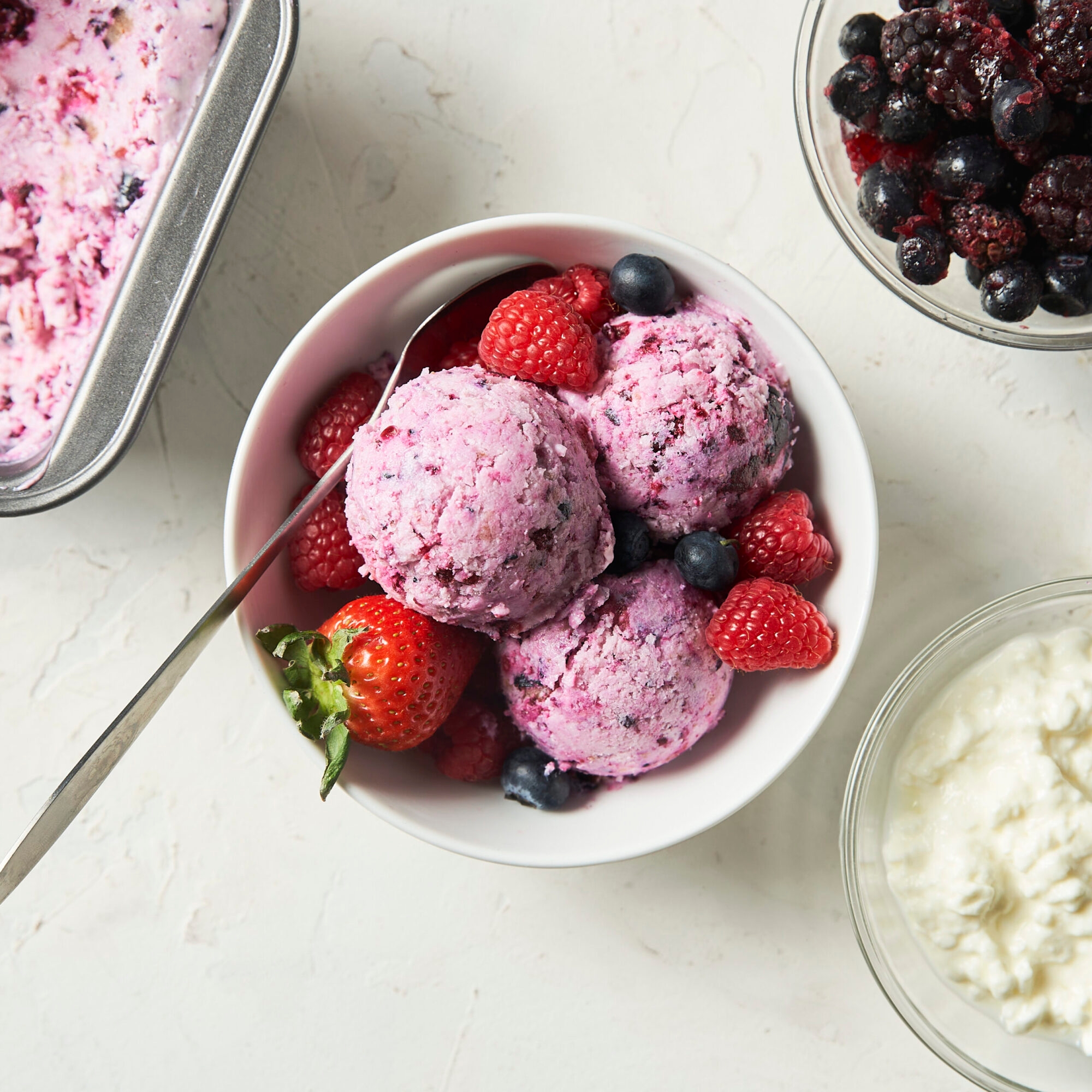 Bowl of berry ice cream with fresh raspberries, strawberries, and blueberries, next to dishes of mixed berries and cream