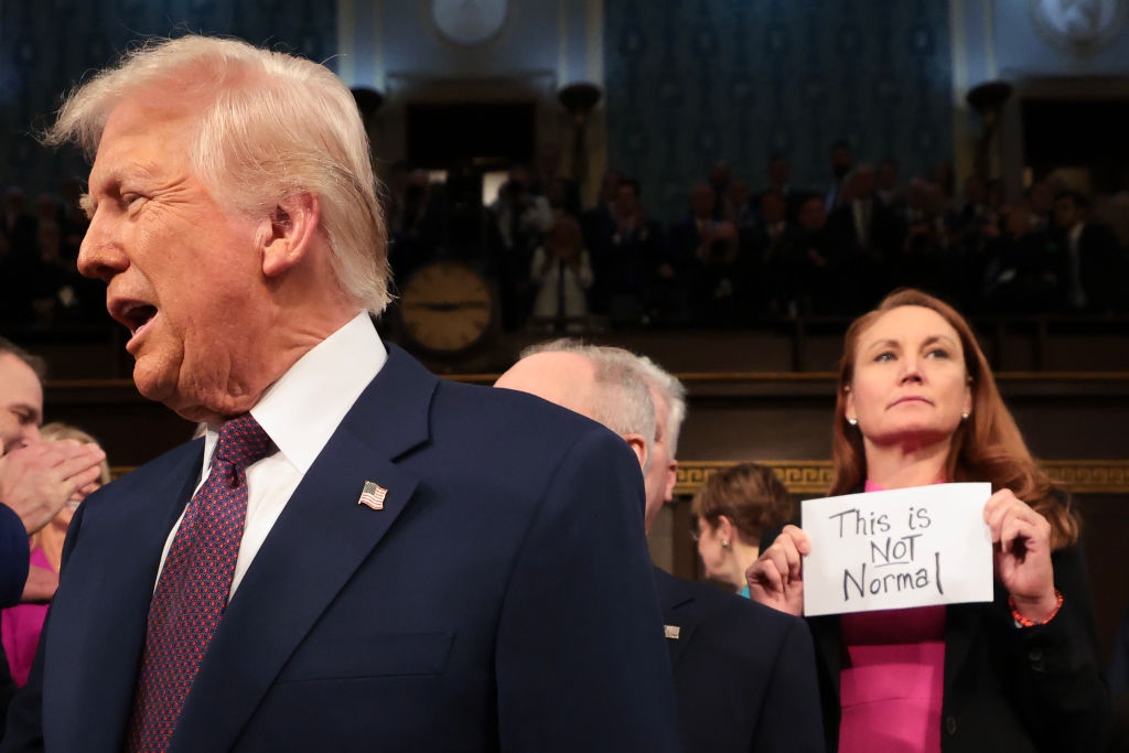 Person in suit appears foreground. Woman holds "This is Not Normal" sign in the background, surrounded by seated audience, suggesting political context