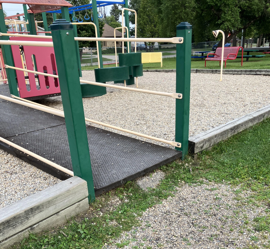 Accessible playground with ramps, a slide, and play structures on a gravel base, surrounded by greenery