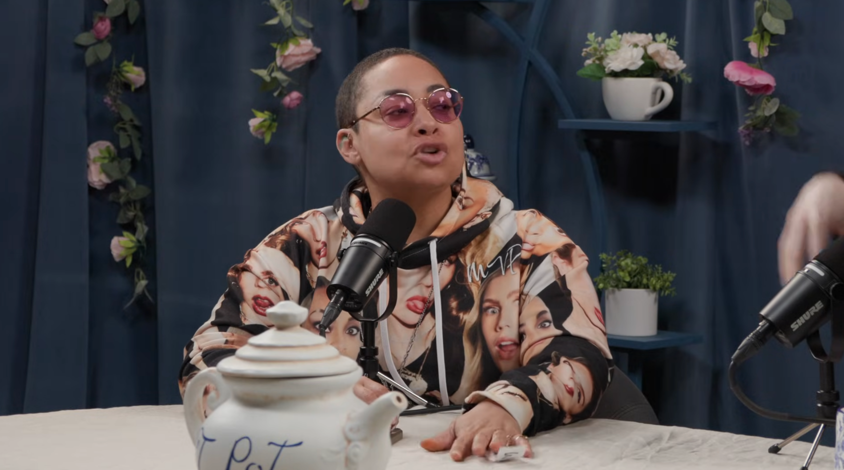 Person wearing a hoodie with face prints sits at a podcast table with microphones and a teapot, speaking animatedly