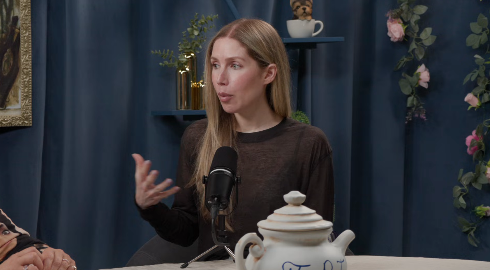 A person sits at a podcast table, speaking into a microphone. They appear engaged in conversation, with a teapot in the foreground