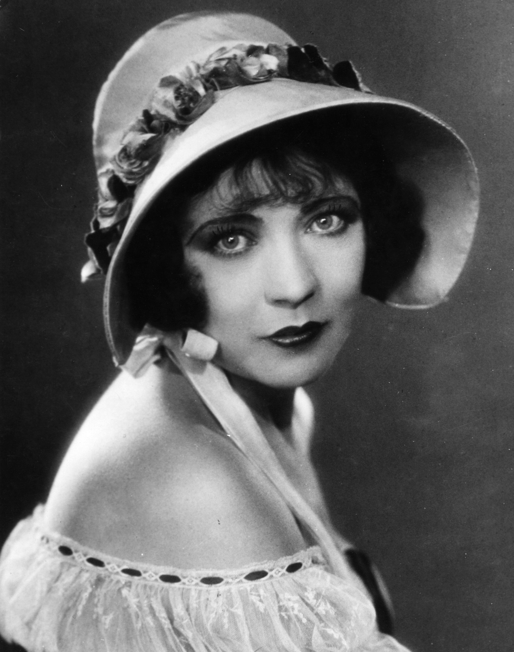 Vintage portrait of a woman with a wide-brimmed hat adorned with flowers, wearing an off-the-shoulder dress, gazing directly at the camera