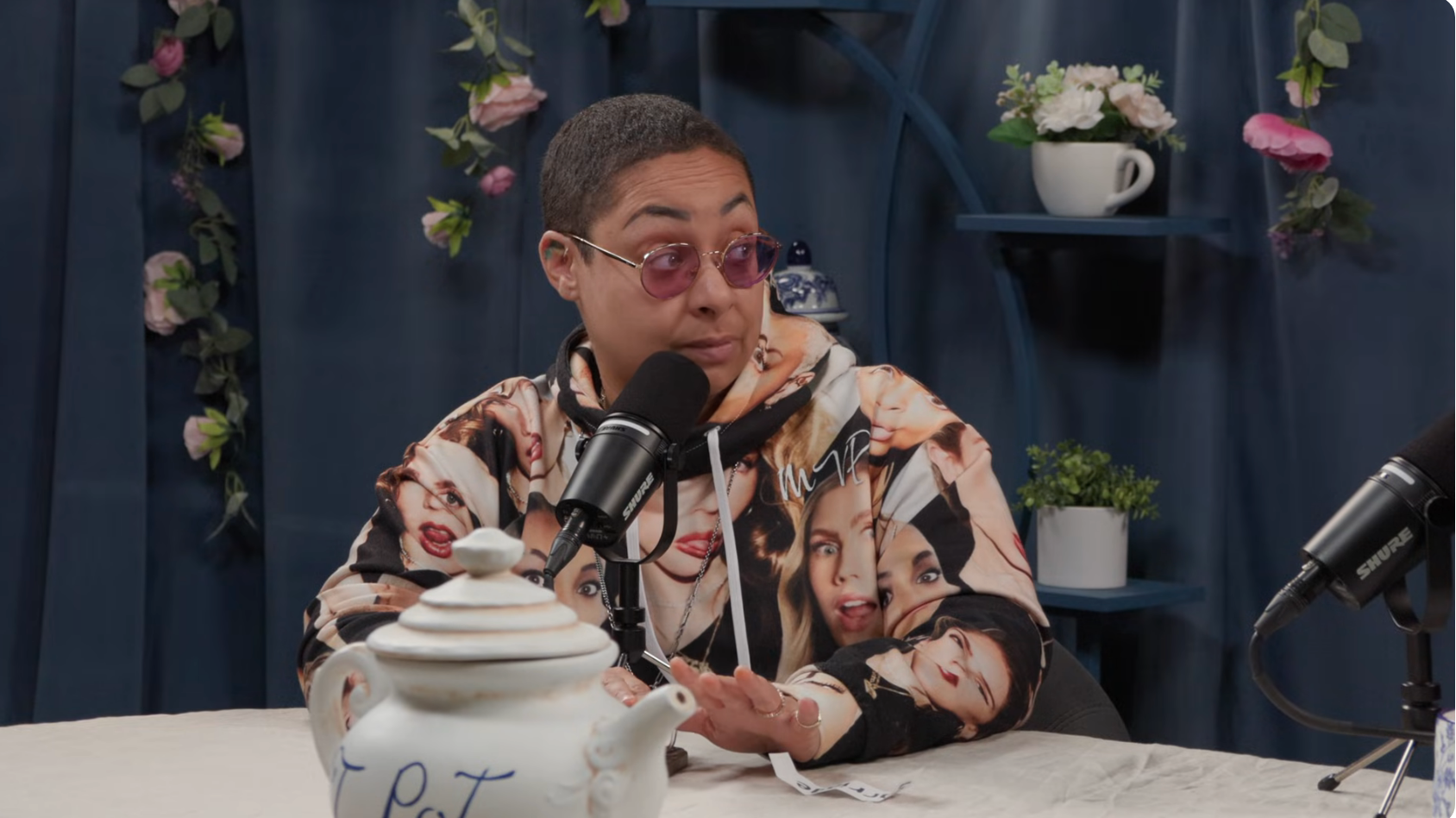 Person speaking into a microphone, wearing a hoodie with various face prints, sitting at a table with a teapot in a studio setting