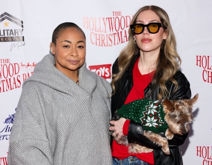 Two people pose on a red carpet. One wears a gray textured sweater; the other sports a black leather jacket, red shirt, and holds a dog in a sweater