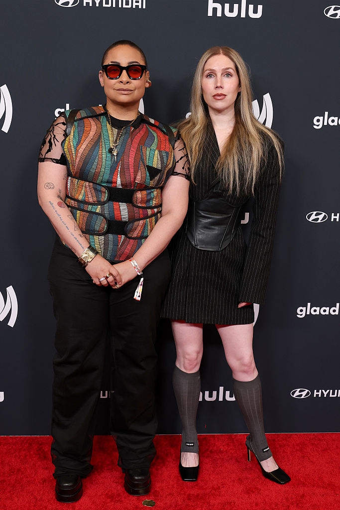 Two people pose on the red carpet at a celebrity event. One wears a colorful patterned top and black pants, the other in a black pinstripe dress and boots