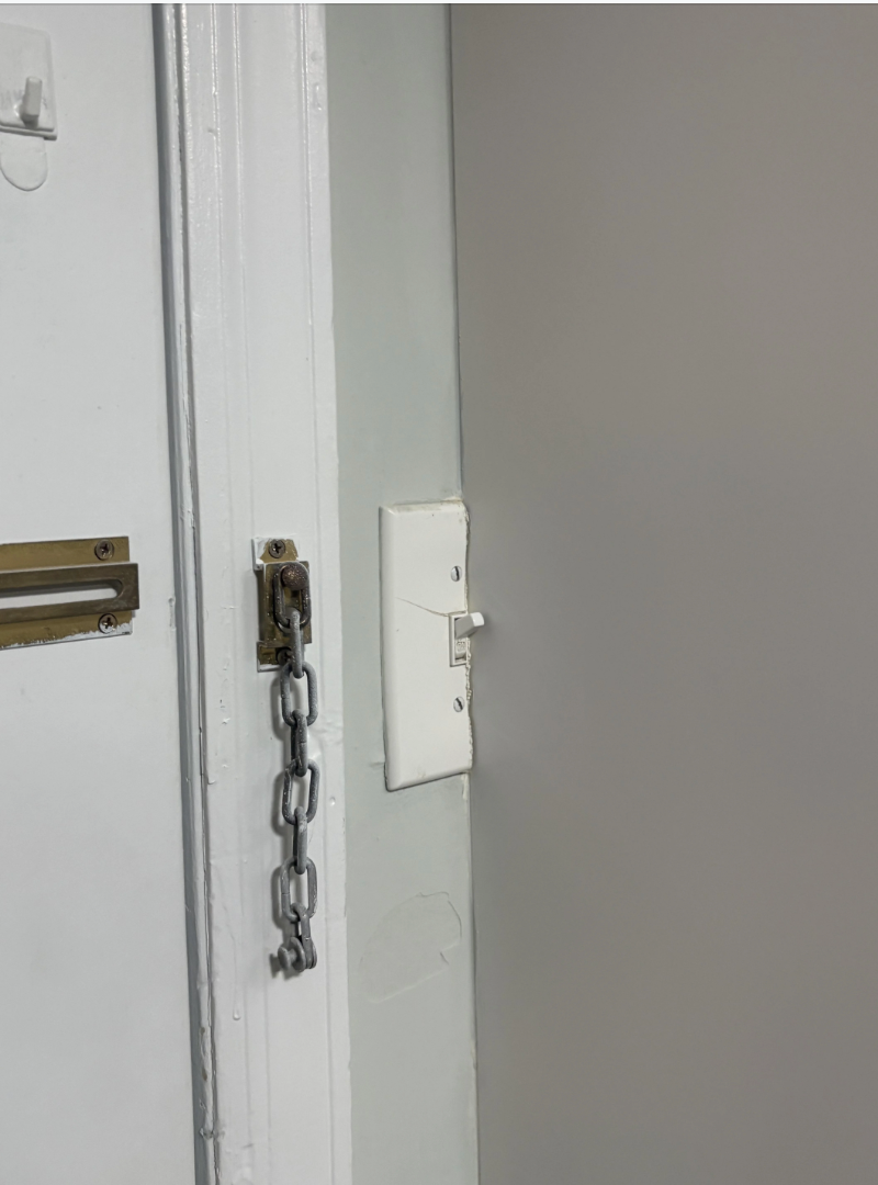 A door chain lock is engaged beside a light switch on a white wall