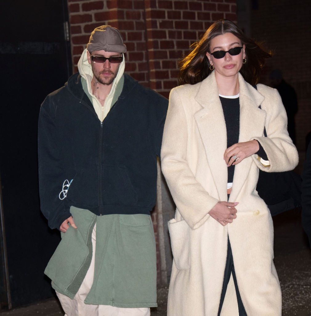Two people walking, both wearing sunglasses. One wears a layered, casual outfit with a hat, the other wears a stylish long coat over a crop top and trousers