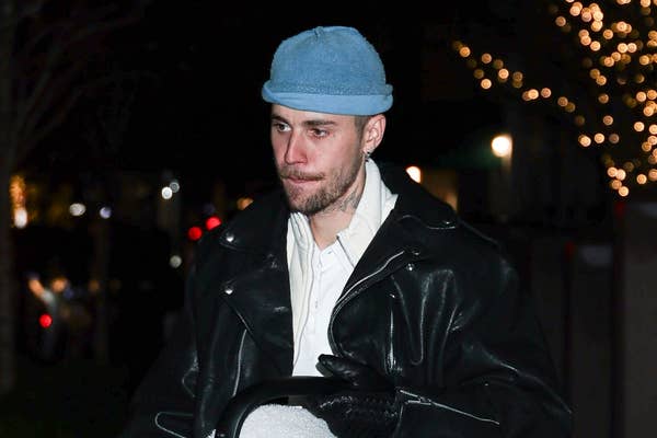 Person in a light hat and leather jacket walking outdoors at night, city lights in the background