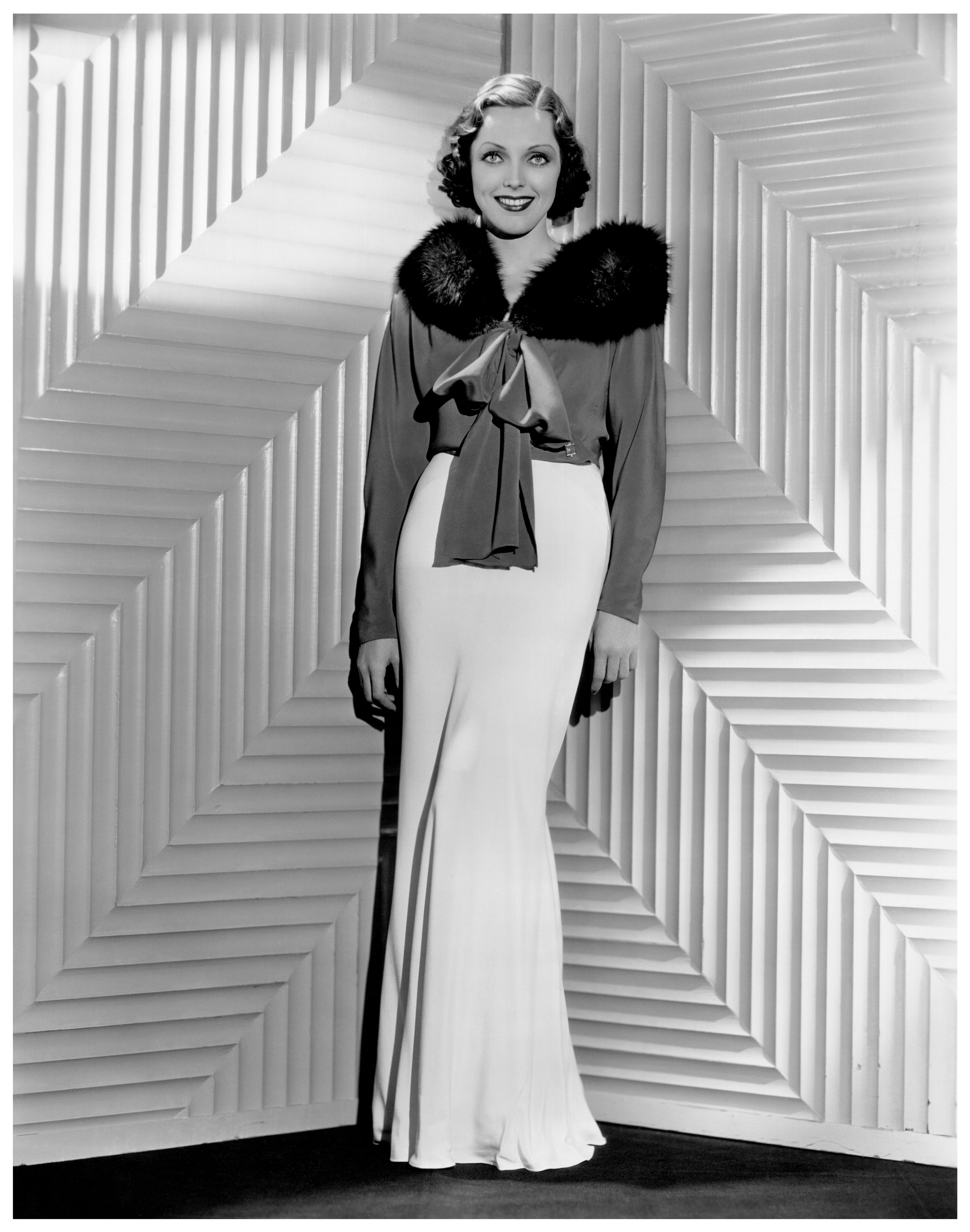 A woman in vintage fashion poses in a floor-length dress with a furry shoulder detail and a bow, standing in front of a geometric patterned backdrop