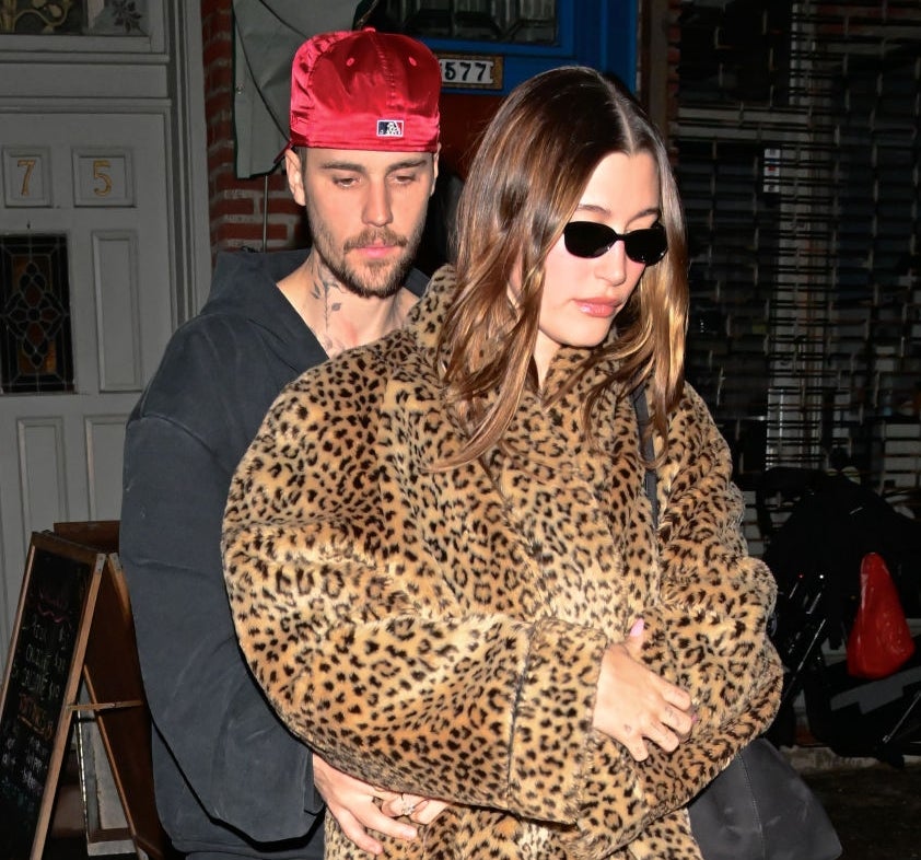 A man in a baseball cap hugs a woman in a leopard print coat and sunglasses on a street at night