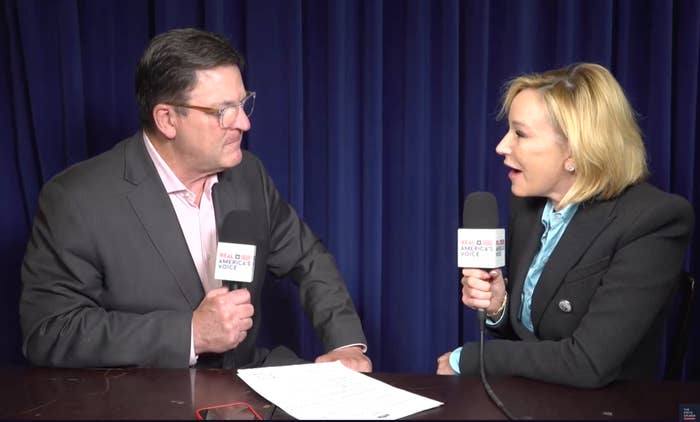 Two people are seated, holding microphones, engaged in an interview in front of a blue curtain background