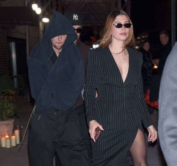 Two people walking; one in a hooded outfit, the other in a pinstriped blazer dress and sunglasses, exuding a chic evening style