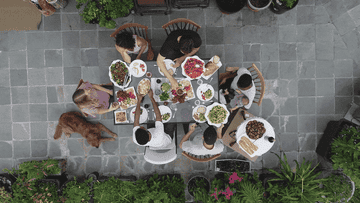 People enjoy a meal at an outdoor table, surrounded by greenery. A dog sits beside them, indicating a relaxed, communal gathering