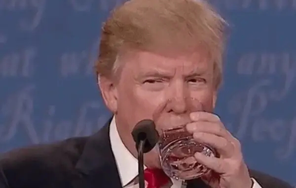 A person in a formal suit is drinking water from a glass behind a microphone on a stage