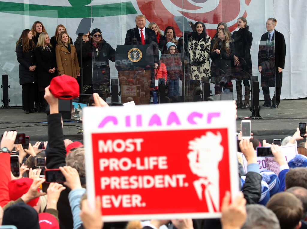 Gruppe mennesker står på scenen med et podium. Et publikum har tegn, man leser & quot; Mest pro-life president noensinne. & Quot; Innstillingen ser ut til å være en offentlig begivenhet