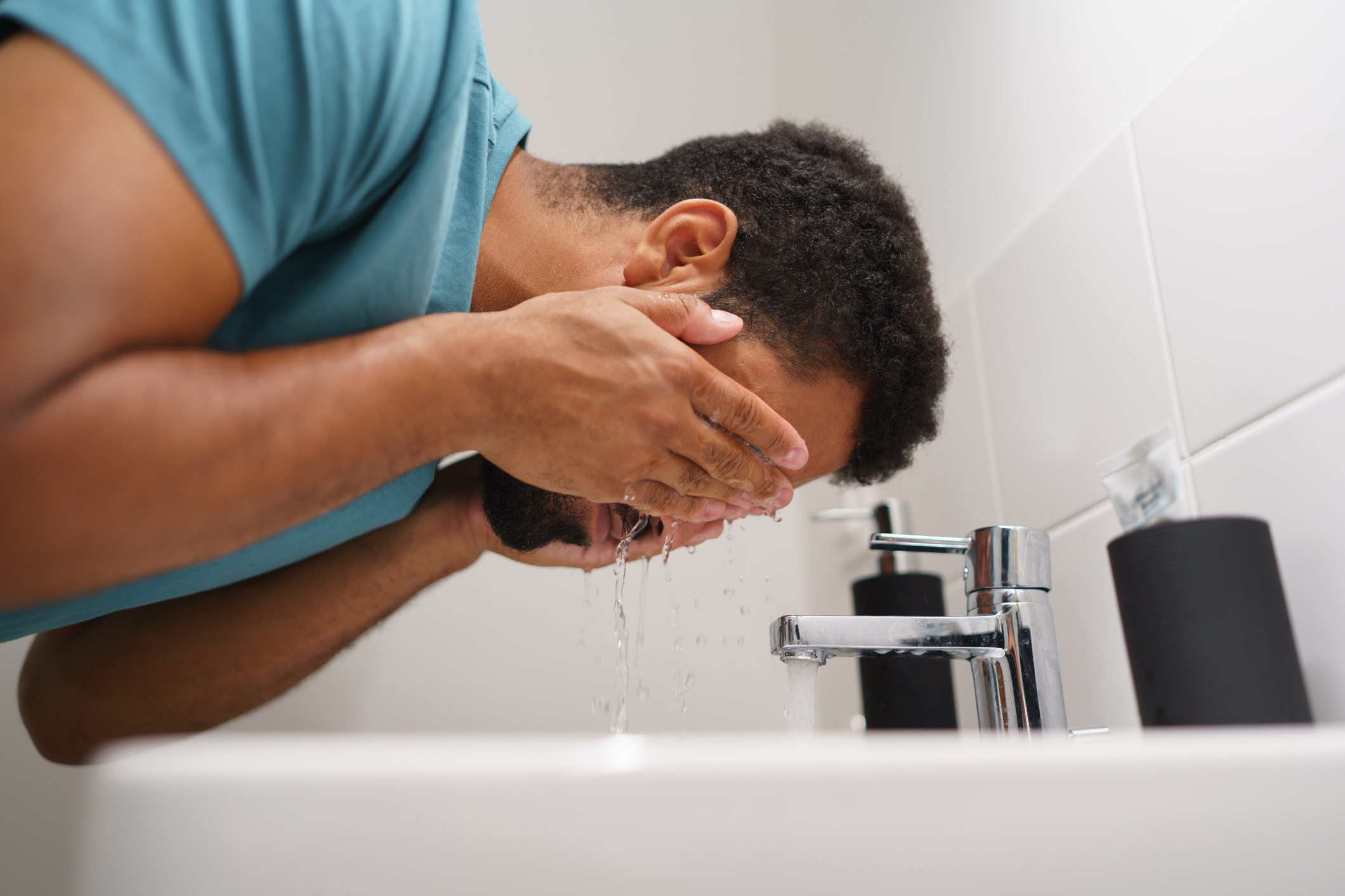 Person washing their face over a sink, water splashing from faucet, hands covering their face