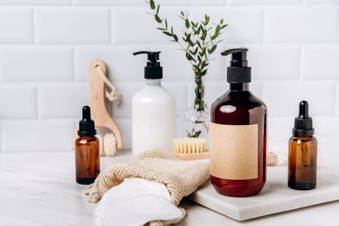 A collection of skincare bottles and accessories on a bathroom counter, including pumps, droppers, and a brush, set against a tiled wall