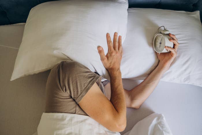 Person in bed shields their head with a pillow, reaching for an alarm clock, suggesting reluctance to wake up