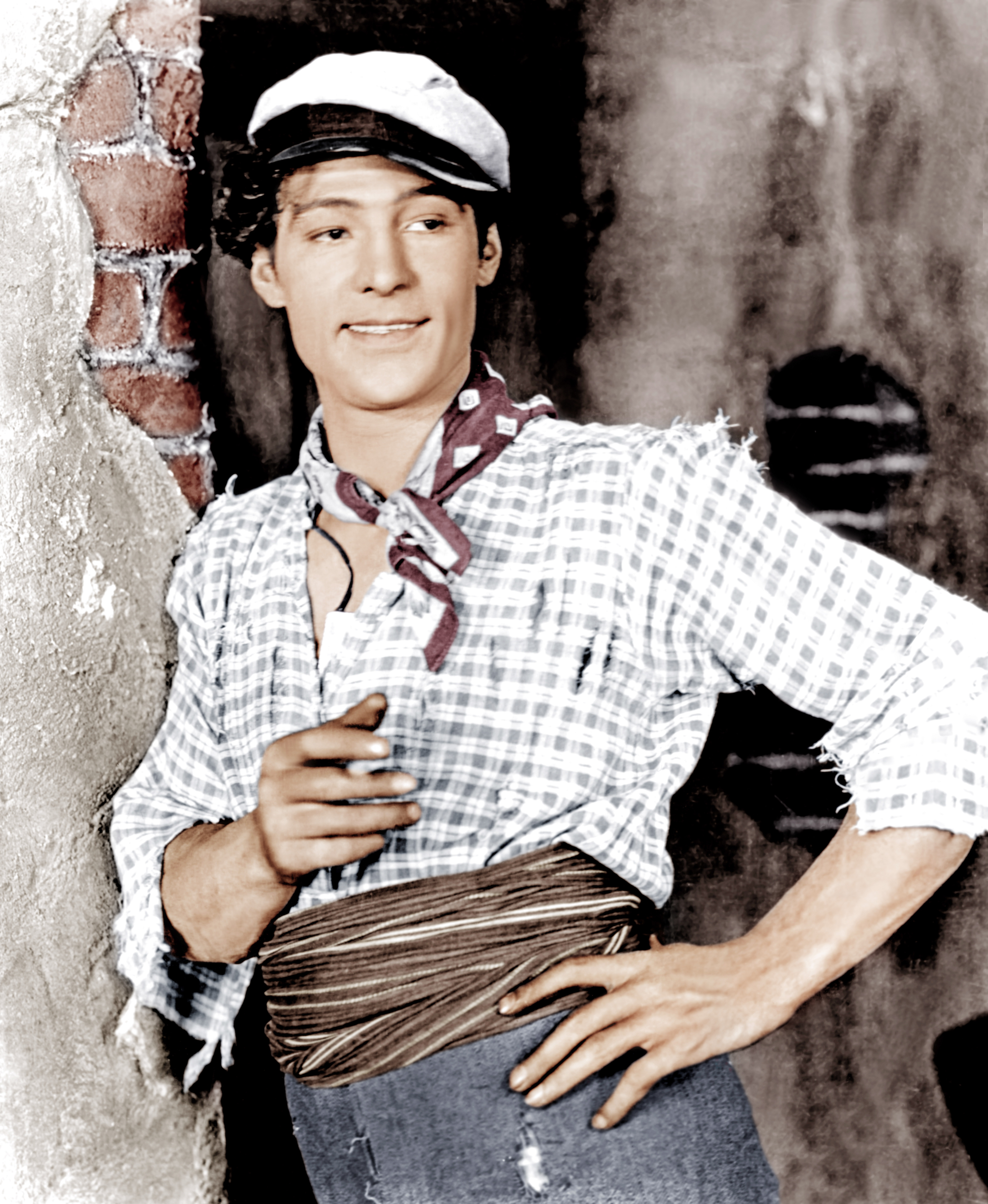 Person in early 20th-century film attire with a checkered shirt, scarf, and cap, leaning against a wall